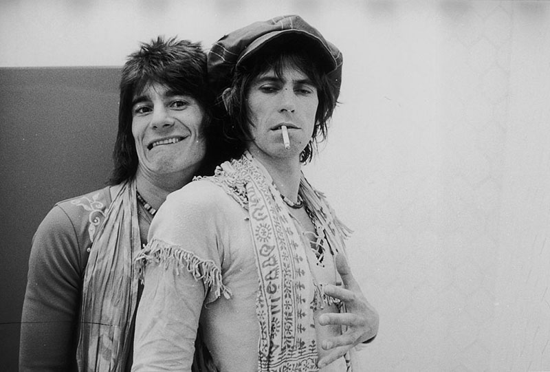 Ronnie Wood wraps stands behind Keith Richards and wraps an arm around him, while Keith holds a cigarette in his mouth and looks down.
