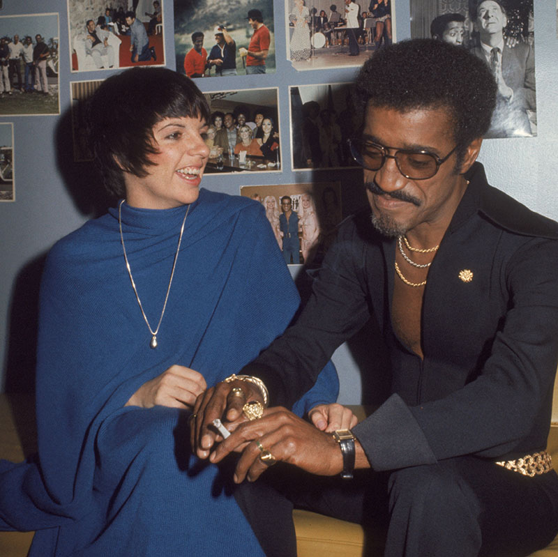Sammy Davis smokes a cigarette while chatting with Liza Minelli in his dressing room, the walls of which are adorn with photos of Sammy throughout his career.