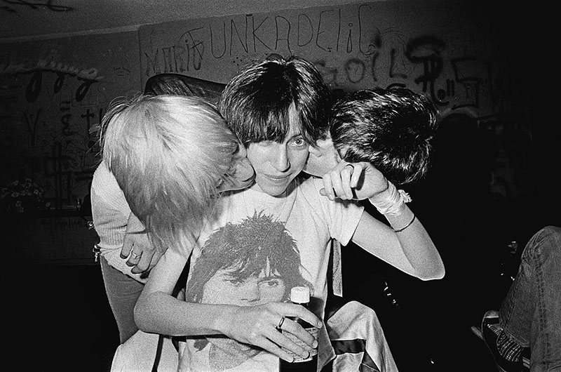 Patti Smith sits in the Whiskey A Go Go while two musicians lean down to kiss each of her cheeks.