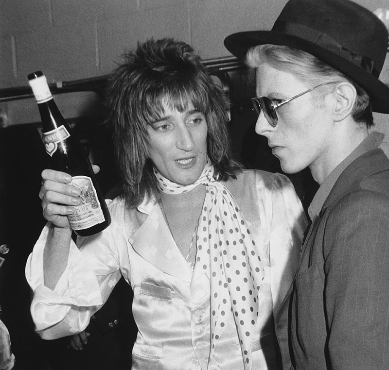 Rod Stewart hold up a bottle of wine while talking to Daivd Bowie, who looks rather ominous with his sunglasses and straightface