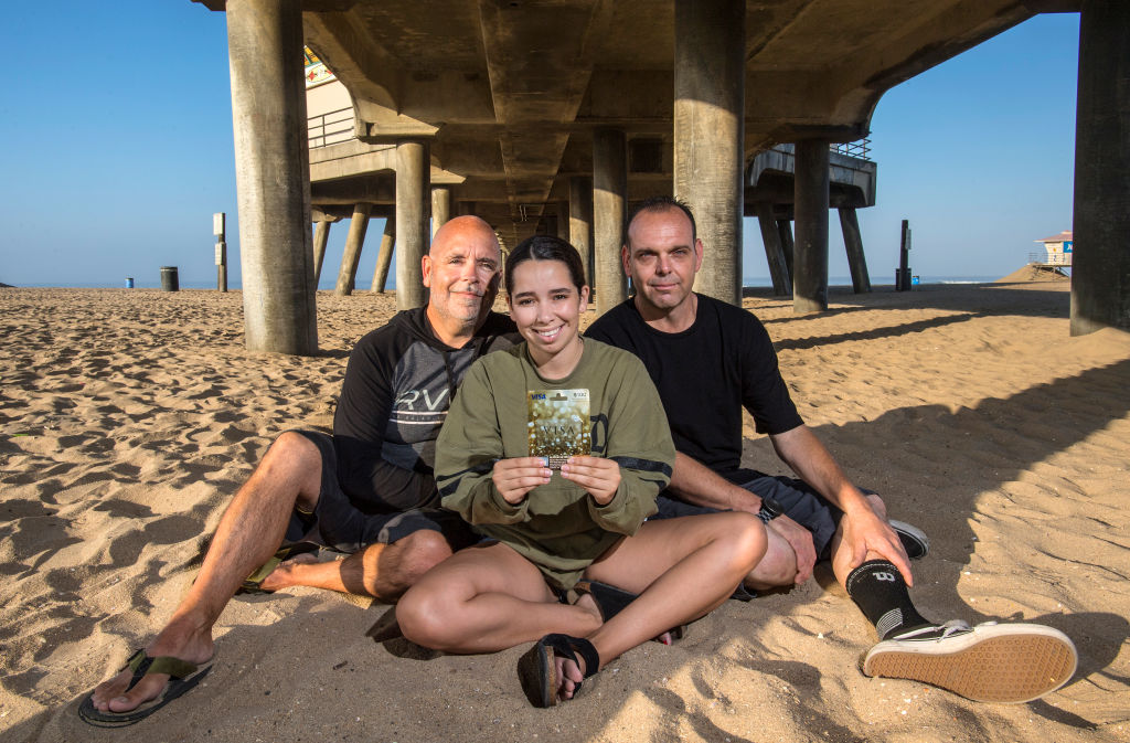 two men and a teen girl sitting on a beach holding a gift card