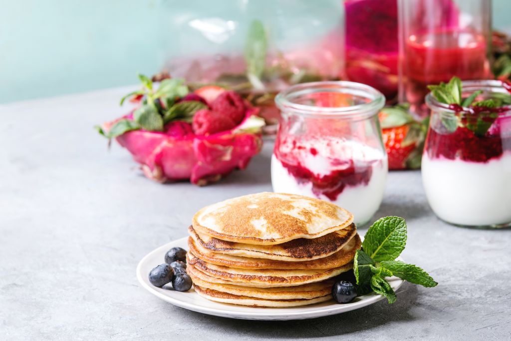 a stack of pancakes on a plate with berries, mint, and glass jars of yogurt