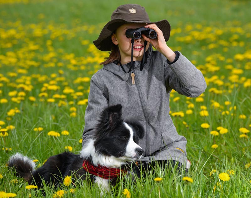 A girl looks through her binaculars while seating in a grass field with her border collie.