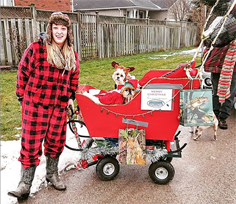 A woman in pjs and cowgirl boots stands next to a santa sleigh on wheels that holds two dressed-up dogs.