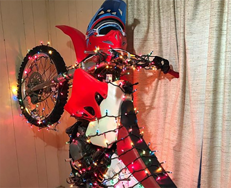 A dirt bike is leaned vertically against a wall and wrapped in Christmas lights.