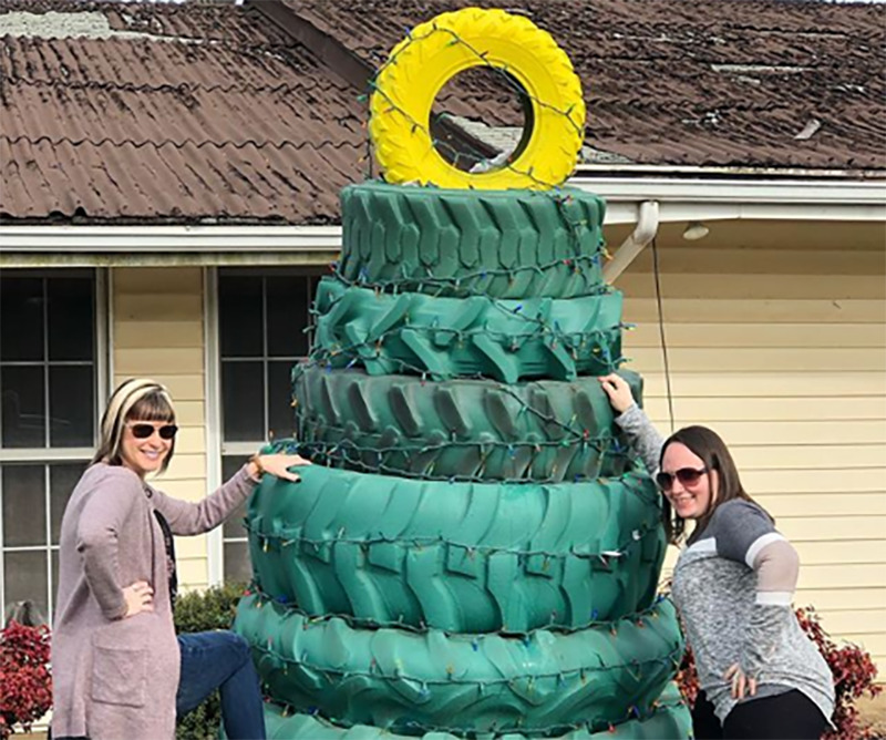 Two woman pose next to a stack of green tires topped with a yellow tire to look like a Christmas tree.