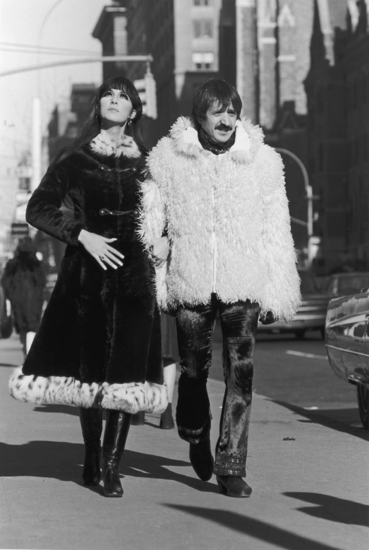 Sonny and Cher walk arm-in-arm down a street in New York wearing large coats.
