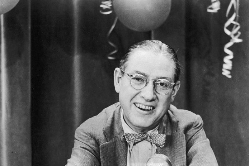 Nash circa 1955: American poet and lyricist Ogden Nash, wearing eyeglasses and a bow tie, smiles while sitting on a television quiz show panel, 1950s.