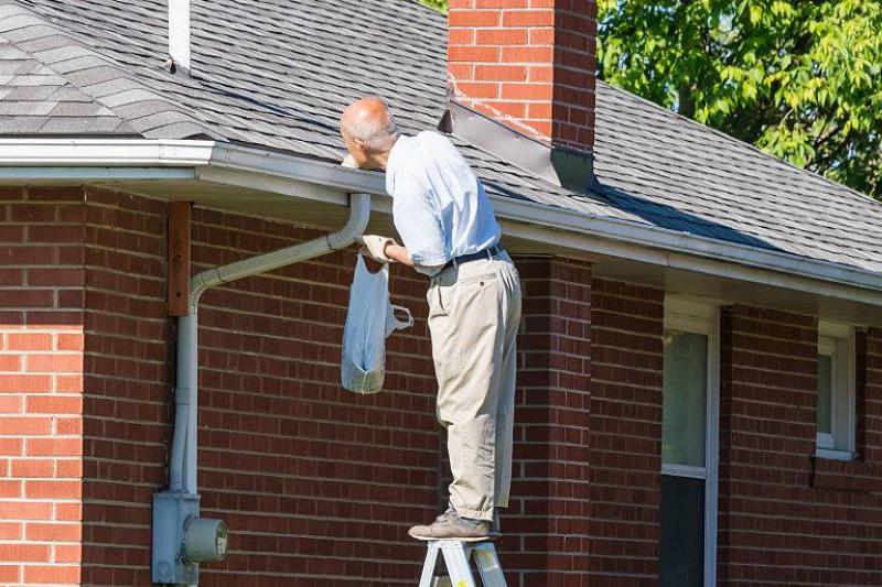 gutters Seniors in Canada: Senior man cleaning a rain gutter on a ladder. Clearing autumn gutter blocked with leaves by hand.
