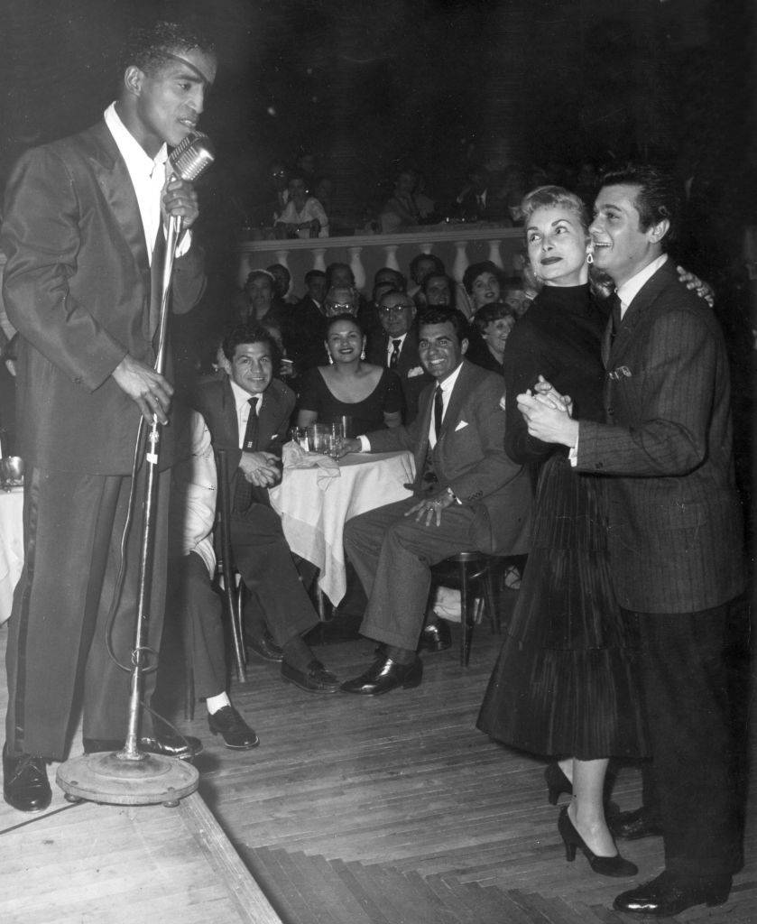American singer and actor Sammy Davis, Jr. (1925 - 1990) sings on stage at a microphone, as married American actors Janet Leigh and Tony Curtis dance