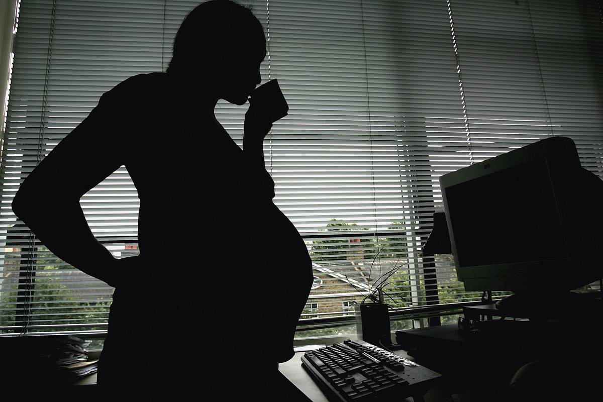 A pregnant woman sips from a mug while standing at a desk.
