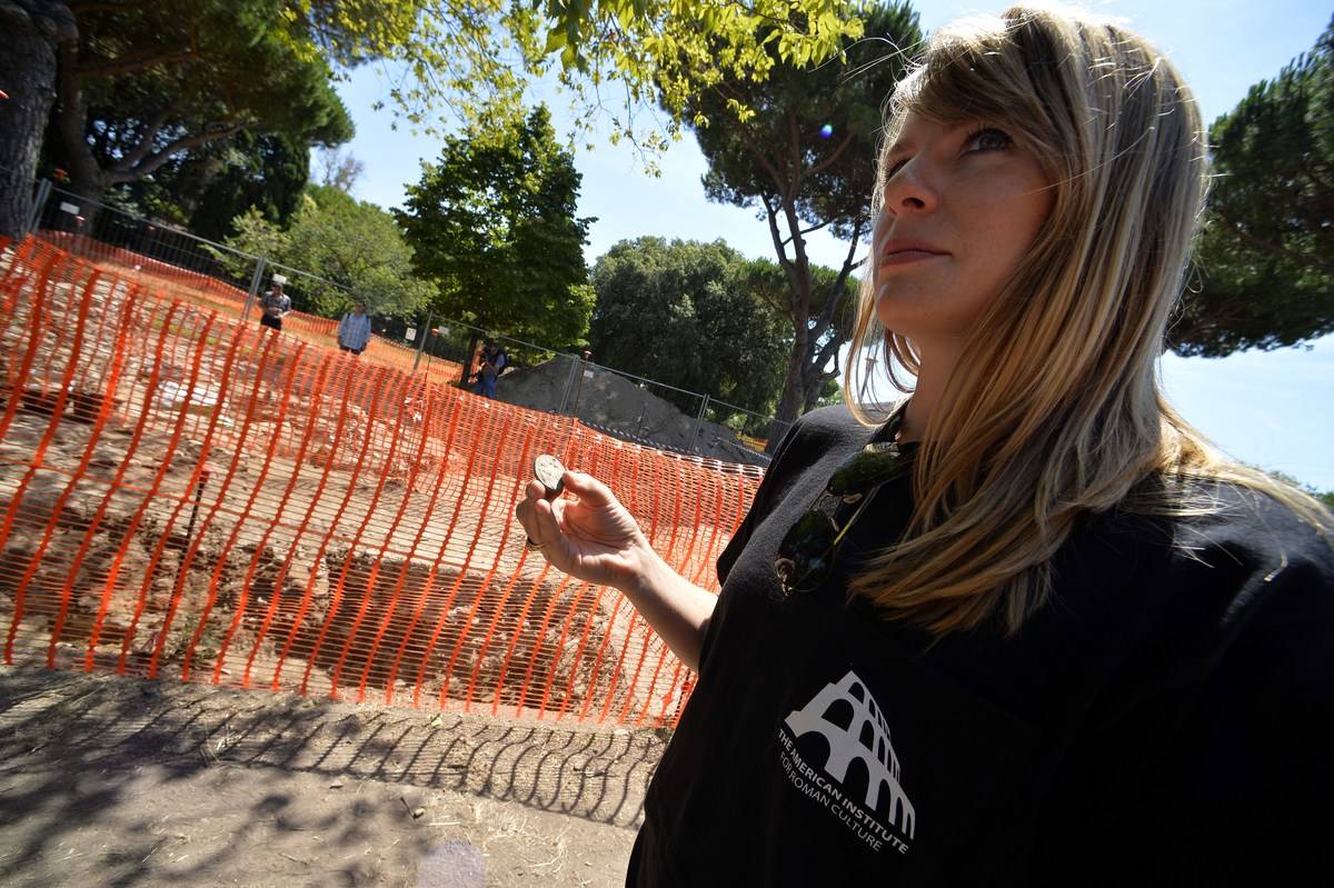 An archaeologist holds a Roman coin in front of a restricted excavation area.