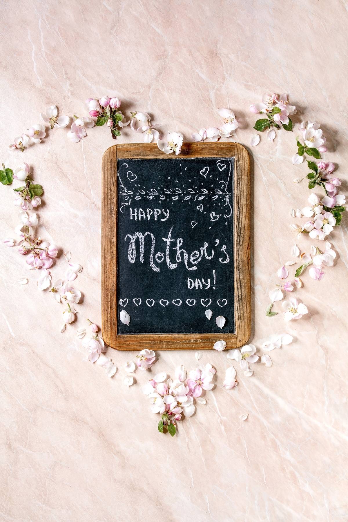 Mother's day greeting card. Vintage chalkboard with handwriting lettering Happy mother's day with spring apple flowers and petals as heart shape over pink marble background. Flat lay, space