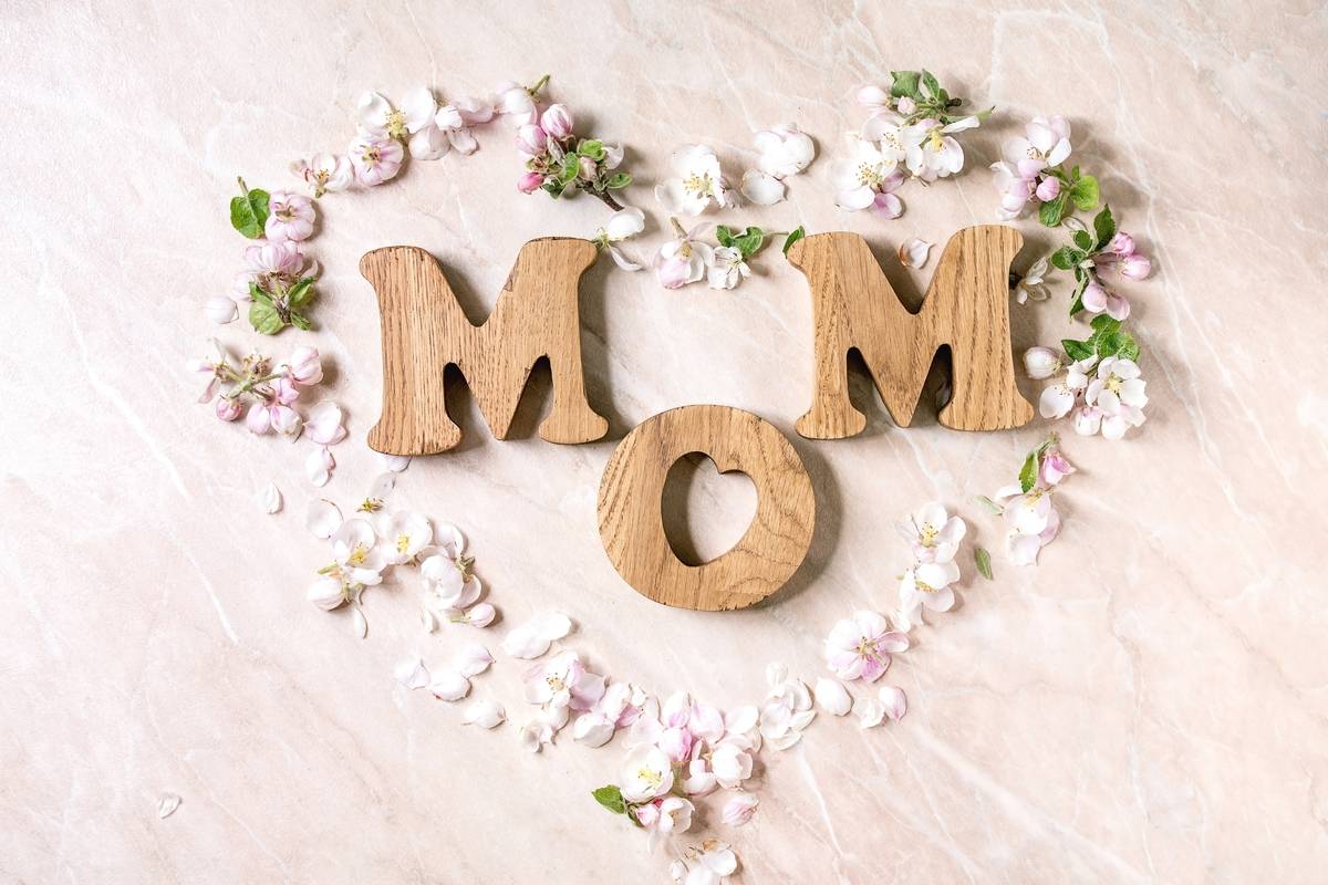 Mother's day greeting card. Wooden letters MOM with spring apple flowers and petals as heart shape over pink marble background. Flat lay, space