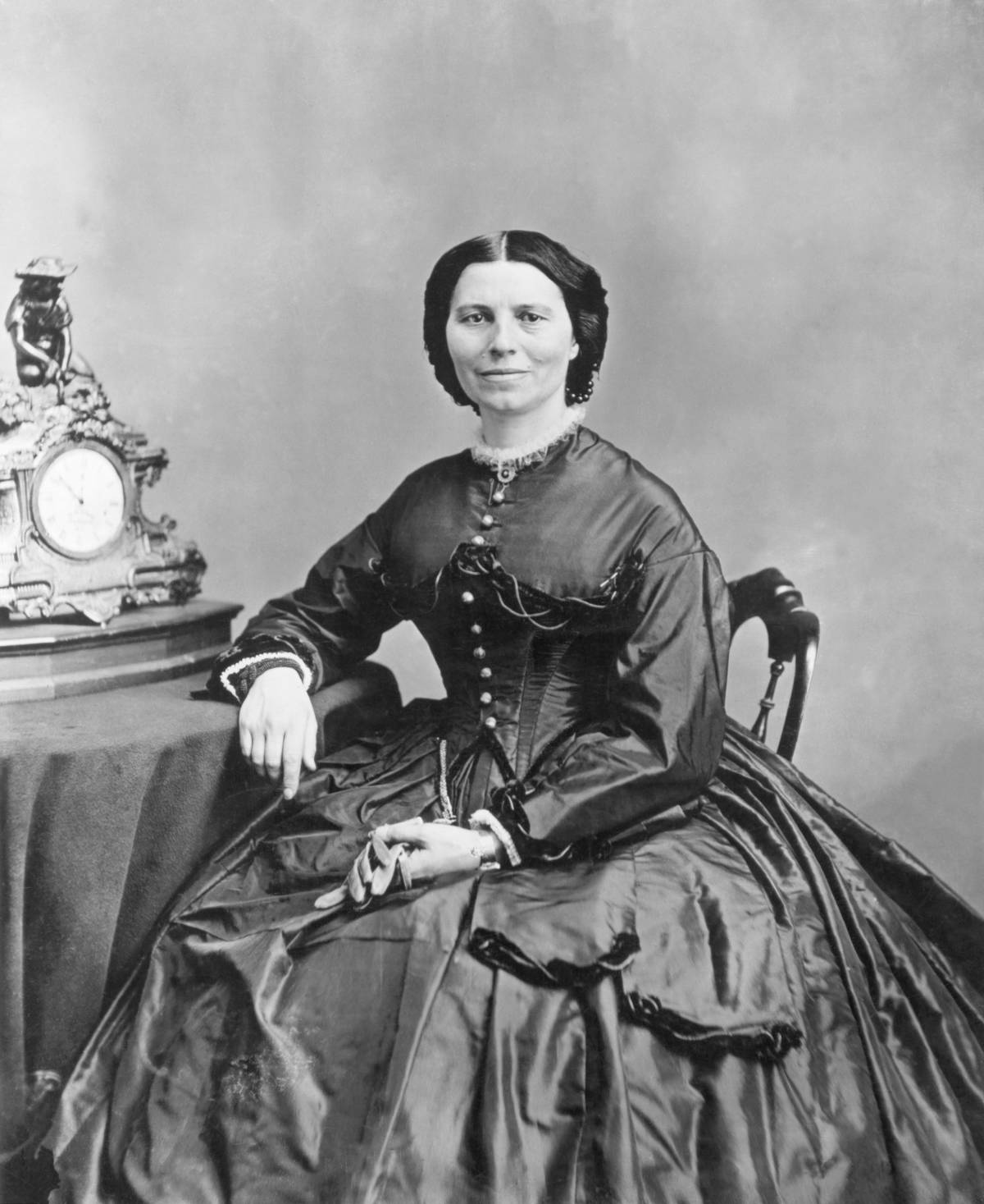 Clara Barton, under President Abraham Lincoln's charge for sending supplies to civil war soldiers, holds a pair of gloves next to a table and clock.