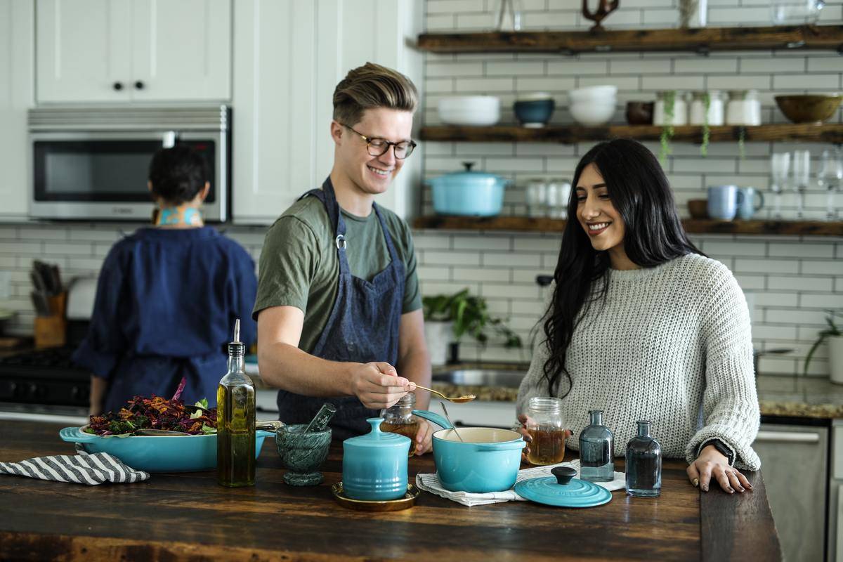 Couple smiles at kitchen island while cooking together, third person at stove