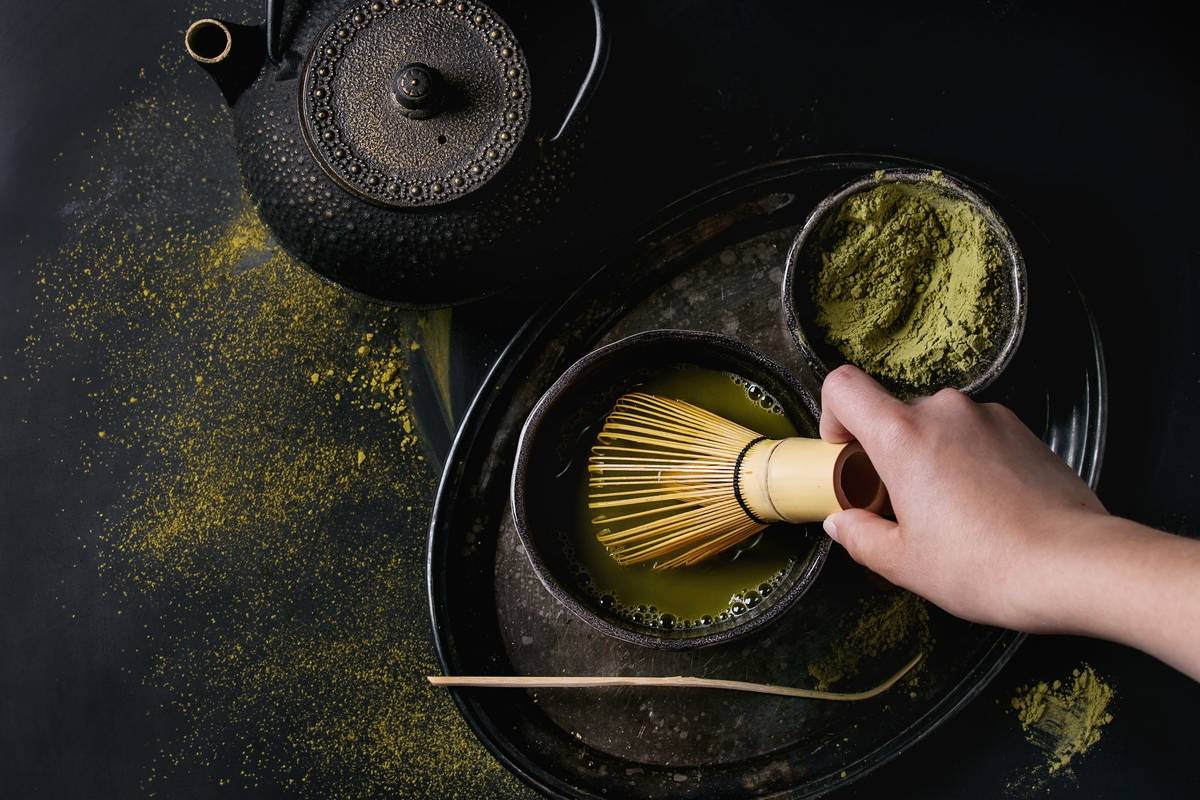 Green tea matcha powder and hot drink in black bowls standing with iron teapot, bamboo traditional tools spoon and whisk in hand in vintage tray over dark metal background