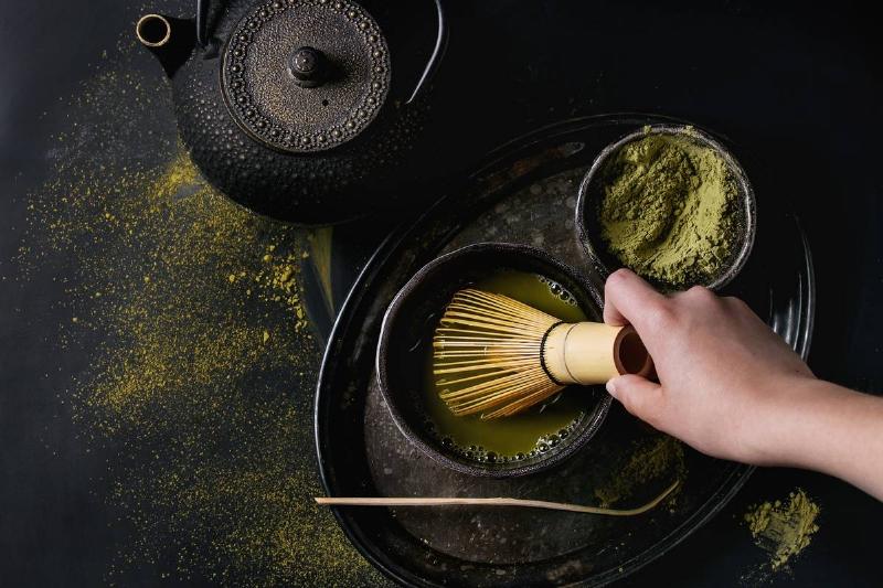 GettyImages-957721924 Green tea matcha powder and hot drink in black bowls standing with iron teapot, bamboo traditional tools spoon and whisk in hand in vintage tray over dark metal background