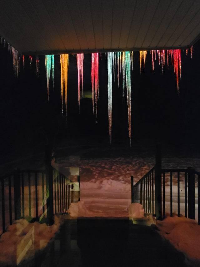 Giant icicles hang off the roof of a home and are lit up in various colors from Christmas lights. 