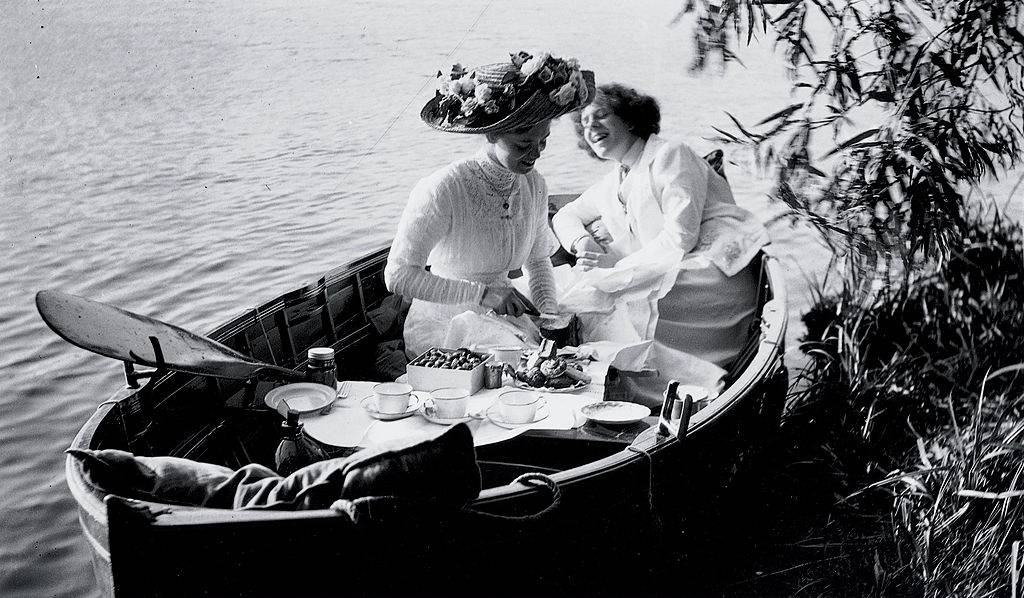 two women in edwardian clothing sitting in a rowboat and having a picnic