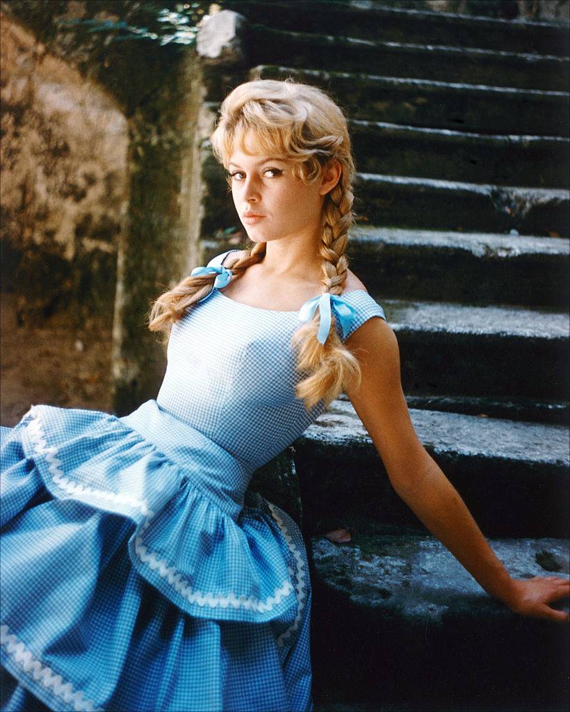 Brigitte Bardot, French actress, model and singer, wearing a blue gingham dress with her hair in plaits, tied with blue bows, sitting in a series of stone steps, 1960