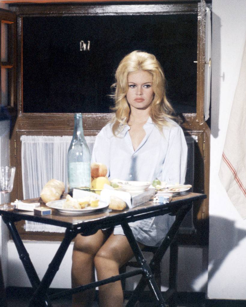 French actress Brigitte Bardot, sitting at a folding table, wearing a man's shirt, circa 1965