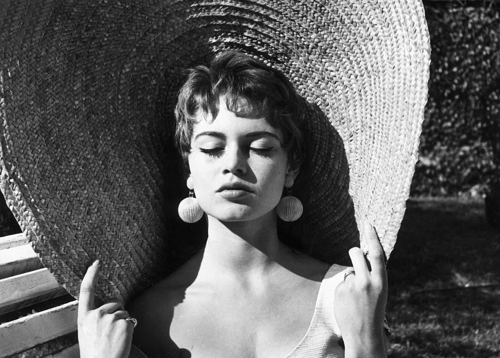Headshot of French actor Brigitte Bardot closing her eyes and holding the rim of her straw hat while basking in the sun outdoors