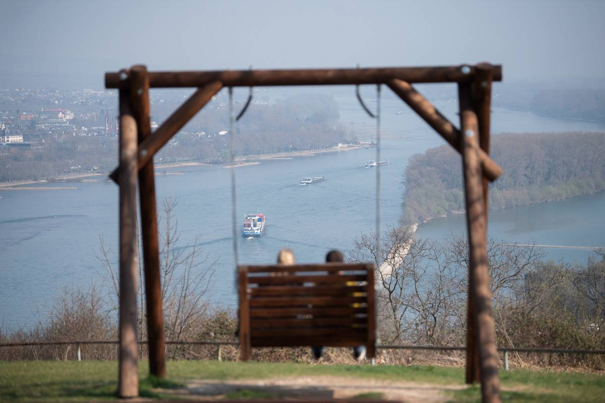 Two tourists are sitting on the Rochusberg in front of the panorama of the Rhine valley. 