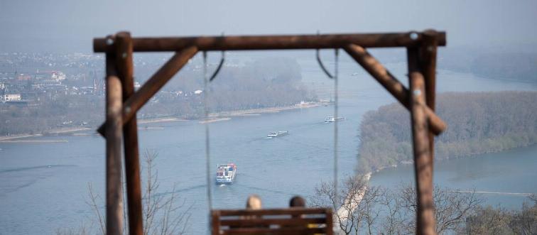 Two tourists are sitting on the Rochusberg in front of the panorama of the Rhine valley.