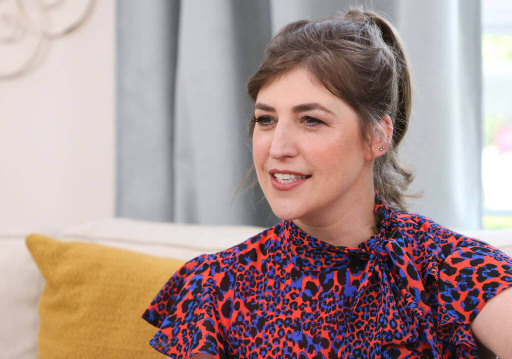 Mayim Bialik sits on couch in interview wearing bright patterned top 