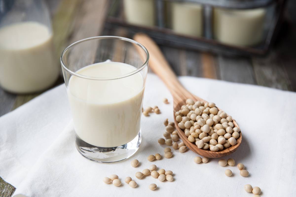 A glass of soy milk next to a wooden spoon with soy beans 