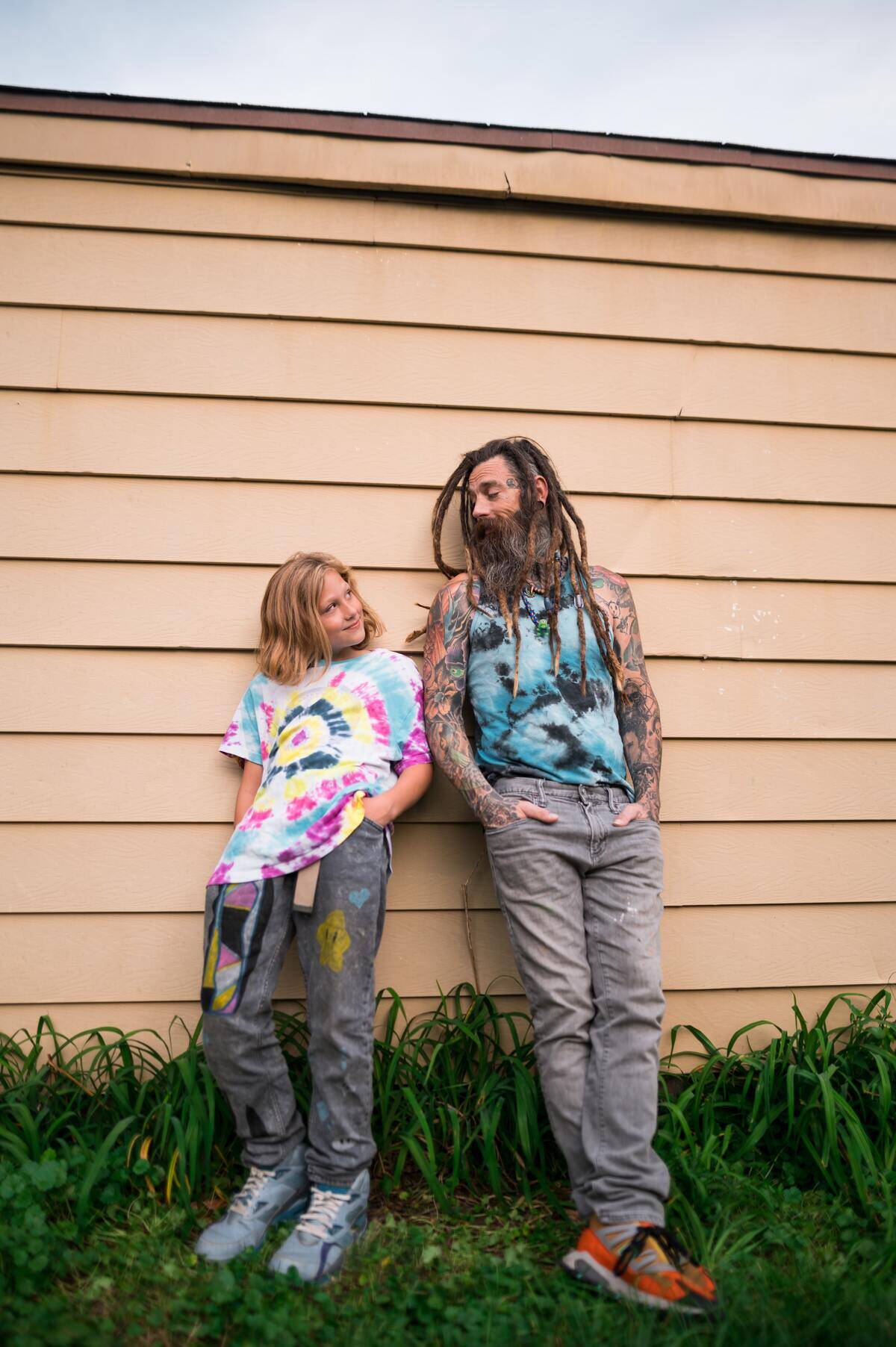 Dad with long dreadlocks leans against house with teen daughter in tie-dye shirt