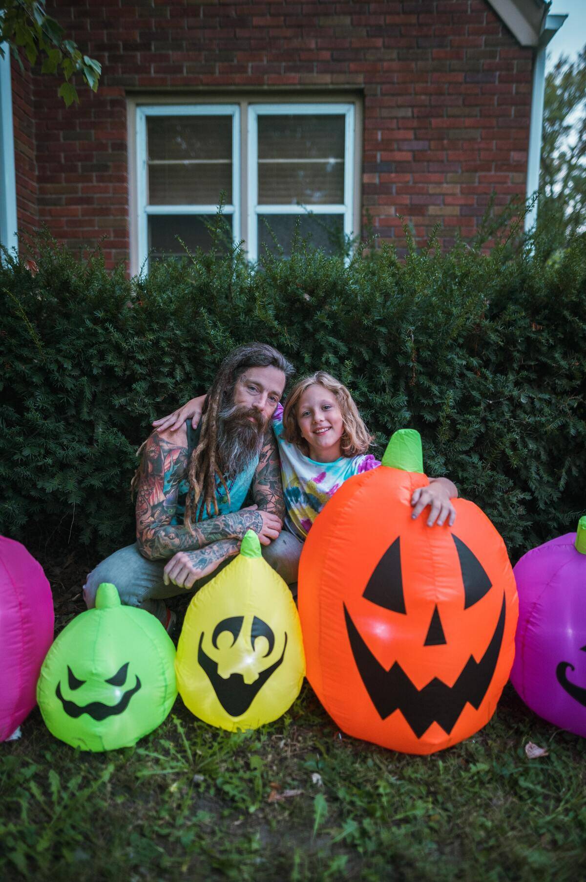 dad with dreadlocks sits next to daughter in tie-dye shirt among colorful lack-o-lantern deocrations for Halloween