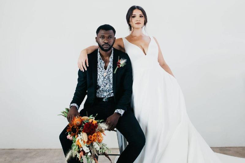 groom sitting and holding flowers with blank expression while bride stands with arm over his shoulder