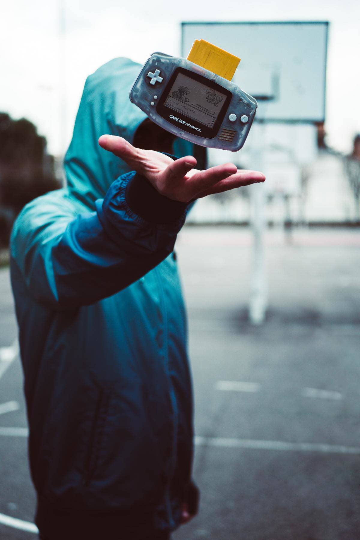 person in hooded jacket throwing up Game Boy Advance with Pokemon Yellow at basketball court