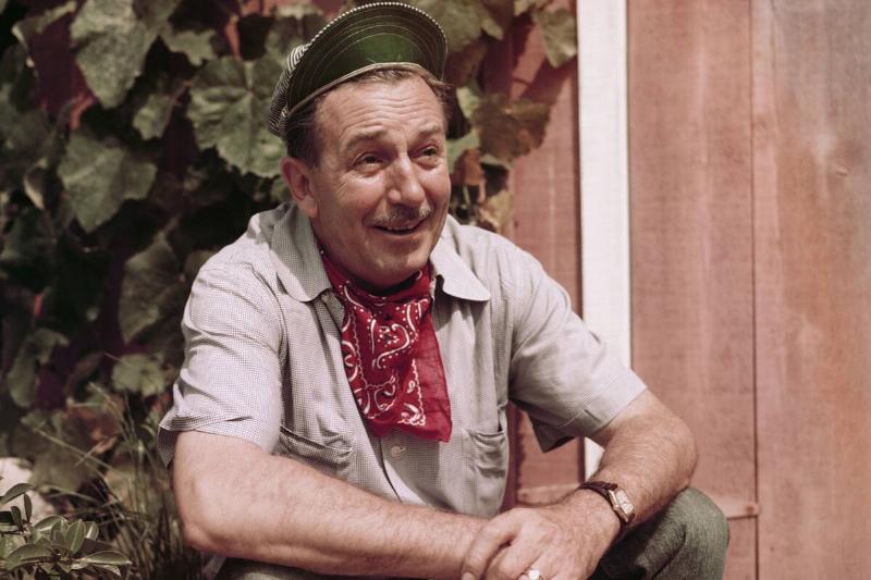 Portrait of American movie producer, artist, and animator Walt Disney  (1901 - 1966) as he sits outdoors beside a wooden door, California, 
the early to mid-1950s. He wears a train engineer's cap and a red 
neckerchief.