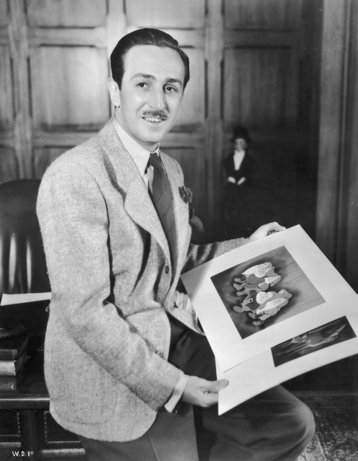 A portrait of American cartoonist and producer Walt Disney (1901 - 1966)  seated on the edge of a desk in an office holding illustrations from 
his animated films 'Snow White and the Seven Dwarfs' and 'Bambi.' 
Animator David Hand directed both films.