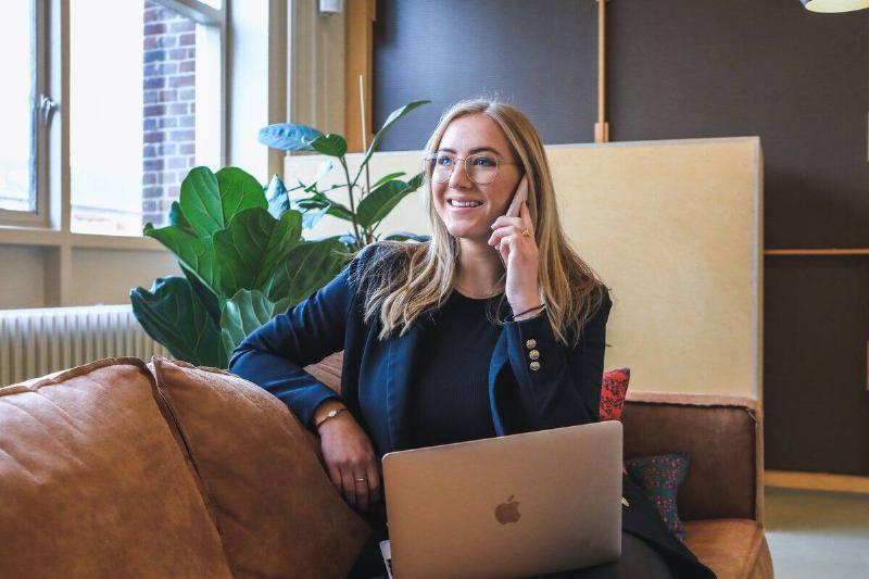 woman in businesswear sitting on couch in front of plant and talking on phone