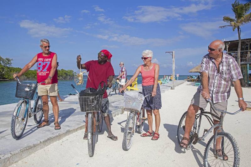 Local black man showing spiny lobster at elderly Western tourists riding bicycles on Caye Caulker.