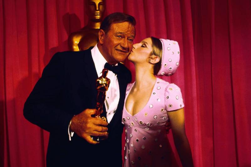 Barbra Streisand Kissing John Wayne in front of a red curtain and a large statue of an Academy Award