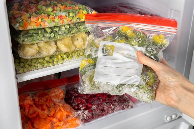 Person off-screen pulling a bag of frozen vegetables from a stocked freezer