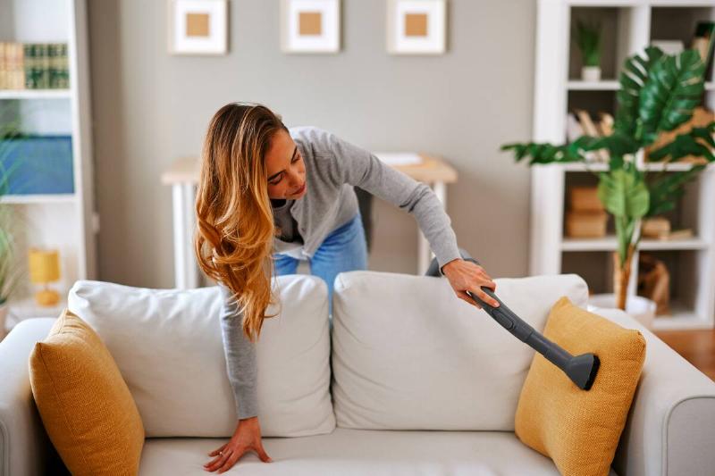 housekeeper-is-vacuuming-a-white-sofa-in-a-modern-2025-05-29-19-12-41-utc Woman vacuuming a white sofa in a modern living room.
