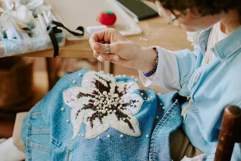 Woman sewing sequins onto denim jacket. 