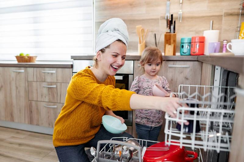 Little girl helps her mom load the dishwasher in the kitchen.