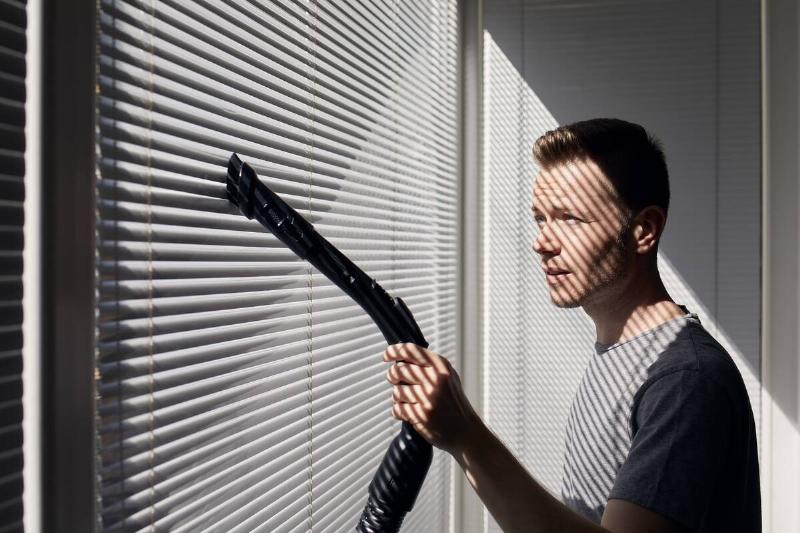 Man cleaning dust from window blind by vacuum cleaner at home.