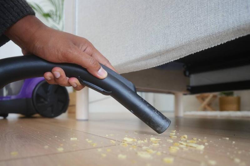 убирать крошки-во время уборки-под диваном-2025-08-24-12-45-26-utc Человек пылесосит крошки с пола под диваном в гостиной.