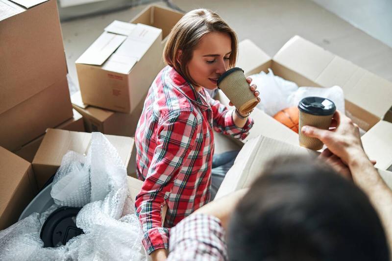 Woman drinking coffee and sitting on the ground, surrounded by moving boxes. 