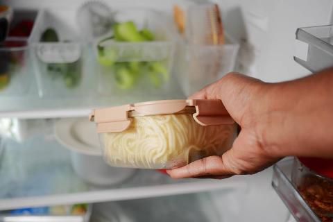 A person holds a container of cooked pasta in a refrigerator filled with food.