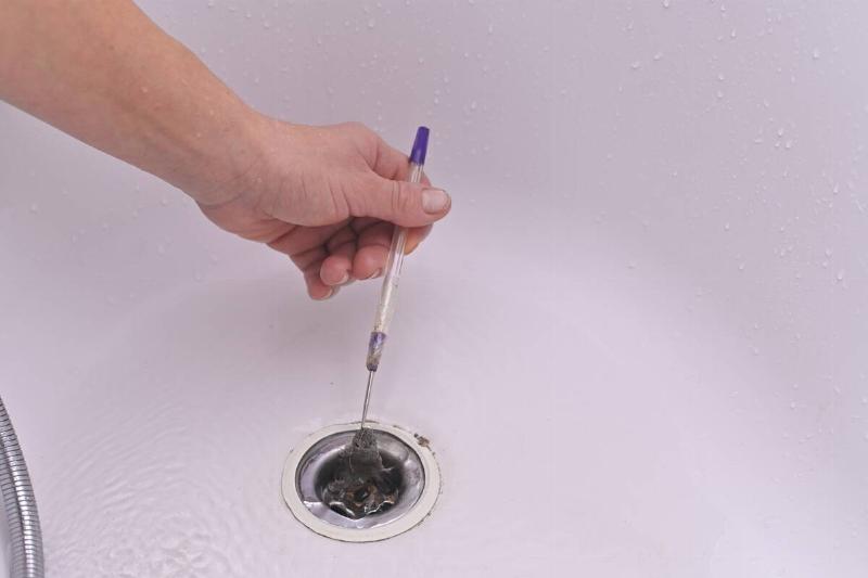 Hand removes hair and dirt from drain using a metal tool.