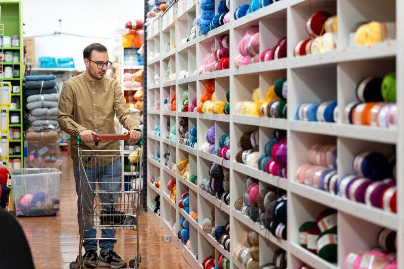 Man pushing shopping cart along aisle filled with various colorful yarn balls and ribbons in a retail haberdashery store.